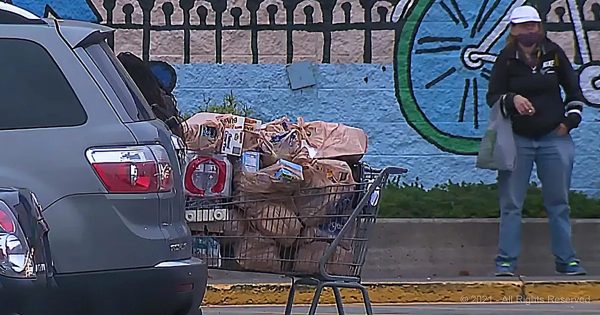 A person stands near a shopping cart filled with bags and groceries, with a mural of a bicycle in the background.
