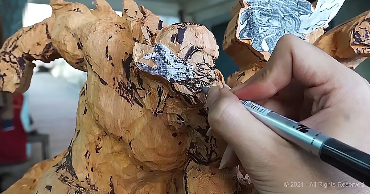 A hand uses a pen to trace a detailed drawing onto a wooden sculpture.