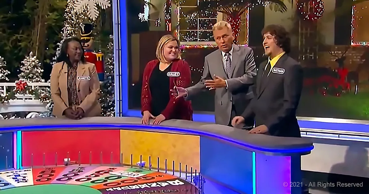 A man in a suit gestures animatedly on a game show set with a festive background.