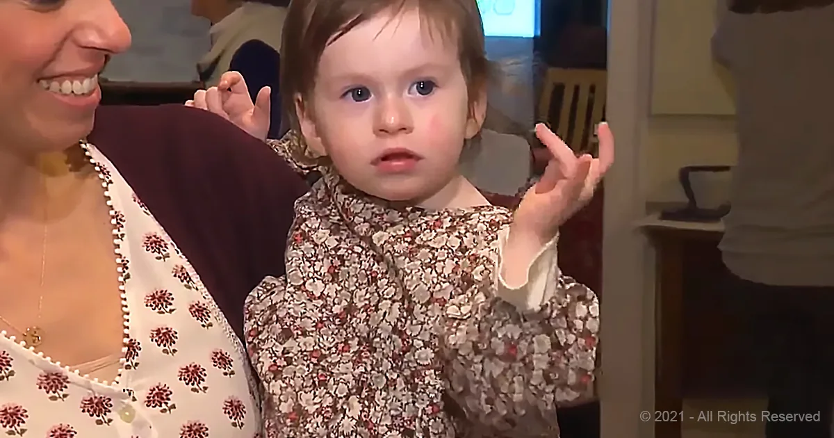 A toddler in a floral dress stands with hands raised, smiling, while a woman in a floral top holds her.
