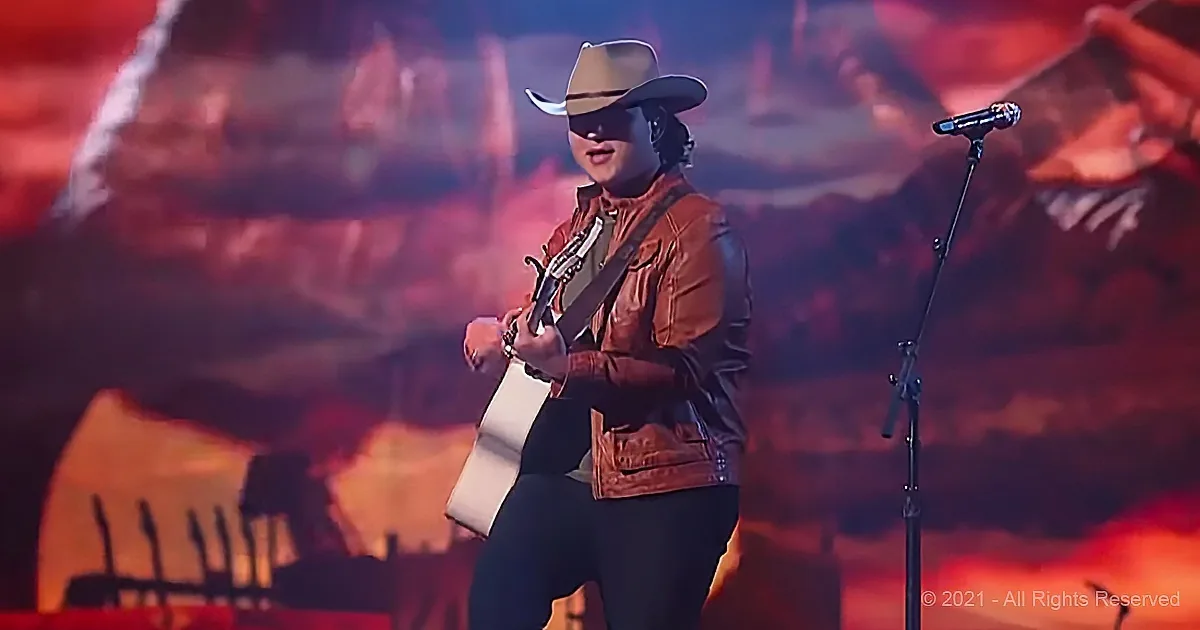 A person in a cowboy hat and leather jacket plays guitar on stage with a microphone and a fiery background.