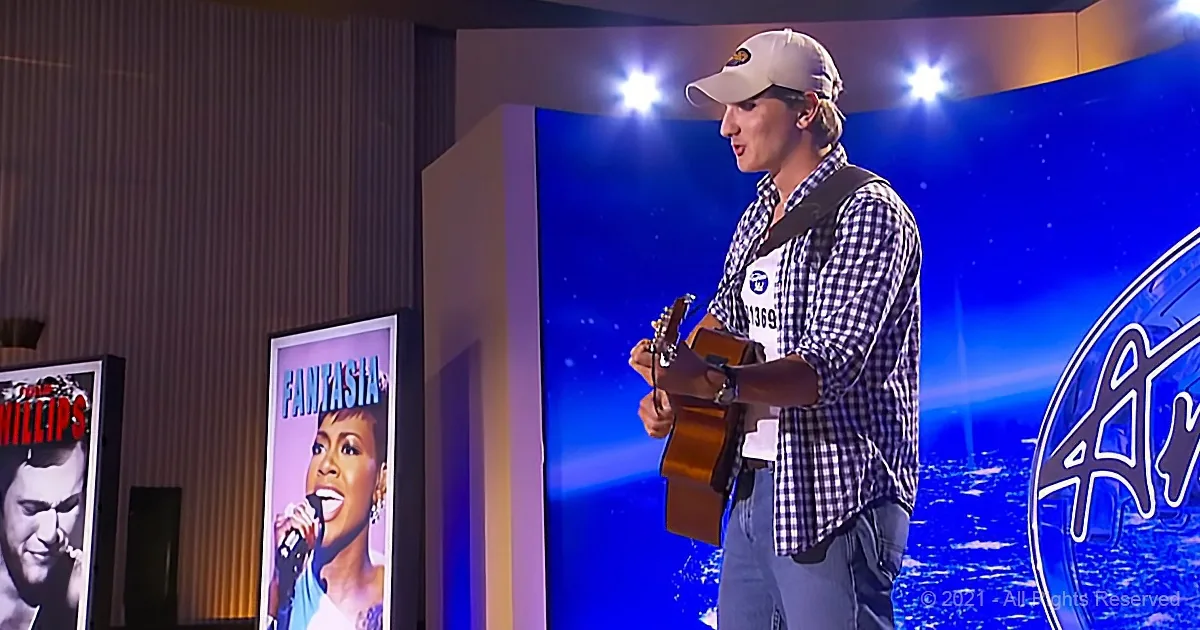 A man in a plaid shirt and cap plays guitar on stage, with posters of performers in the background.