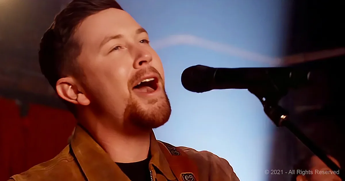 A man with a beard sings into a microphone, wearing a brown jacket and a black shirt.