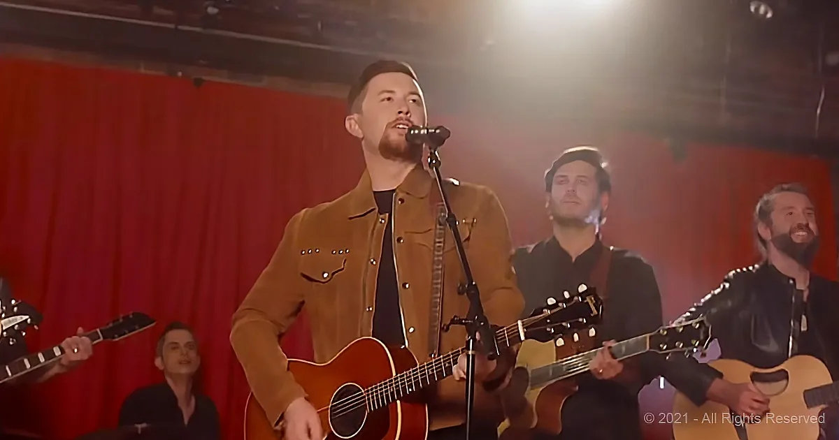 A man in a brown jacket sings into a microphone while playing an acoustic guitar, surrounded by other musicians.