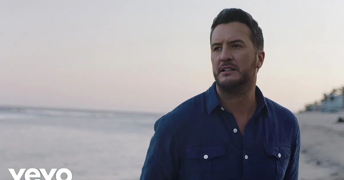 A man with a beard and short hair, wearing a blue shirt, is singing with his eyes closed.