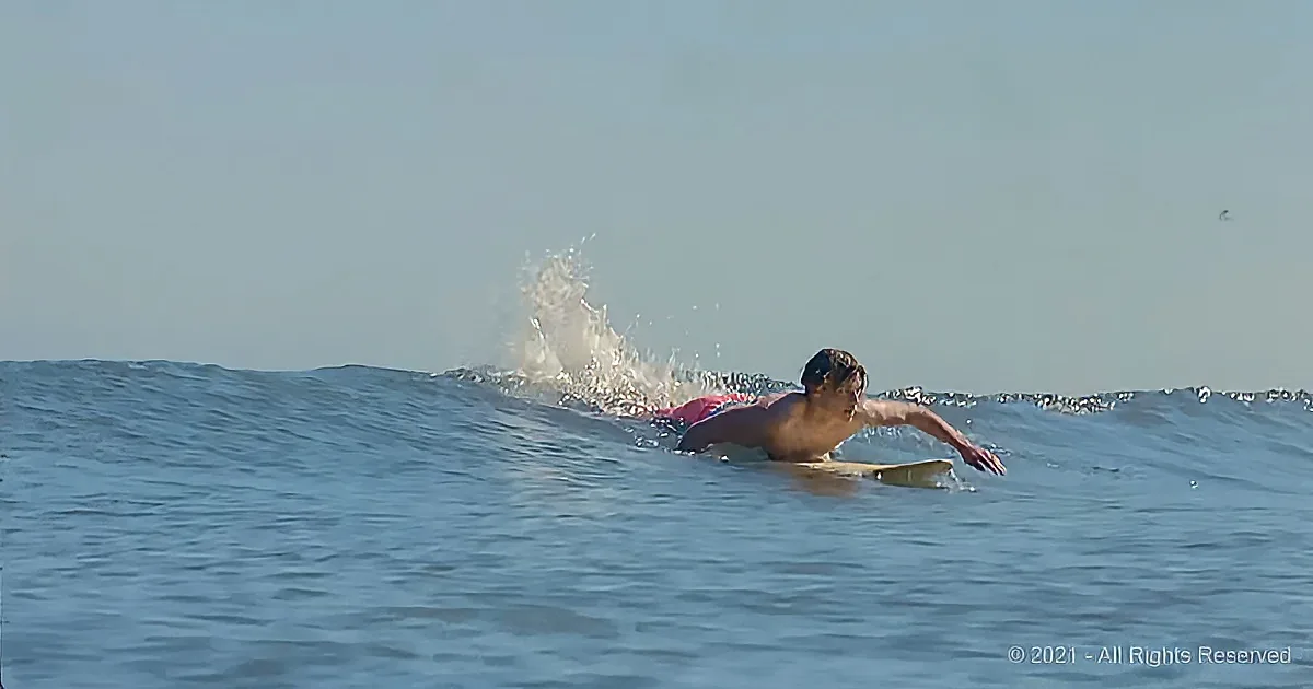 A surfer paddles on a wave, arms extended, water splashing around.