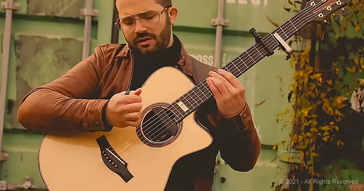 A man in a brown jacket plays an acoustic guitar outdoors, with a green wall and foliage in the background.