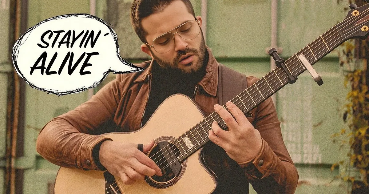 A man in a brown jacket plays an acoustic guitar, wearing glasses and a beard, with a green background.