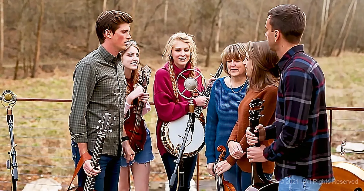 A group of people with musical instruments, including a banjo and violins, gather around a microphone in a rustic outdoor...