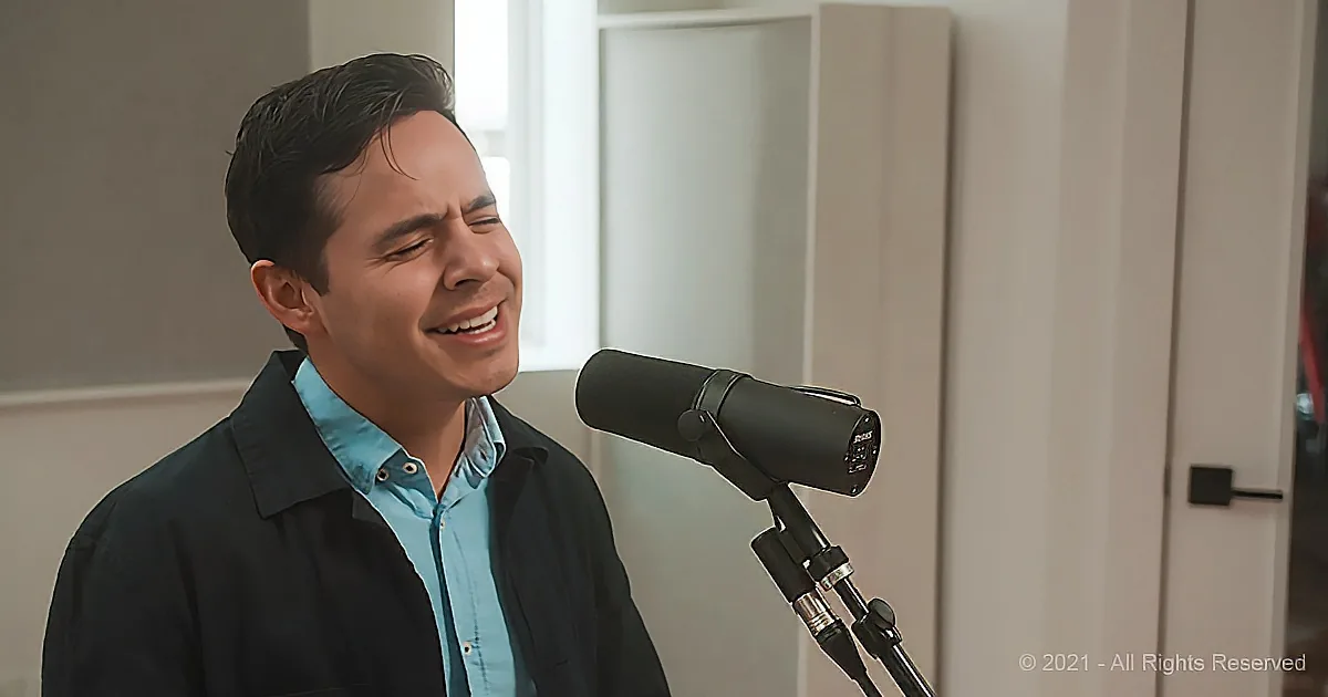 A man in a blue shirt sings into a microphone in a room with light-colored walls.