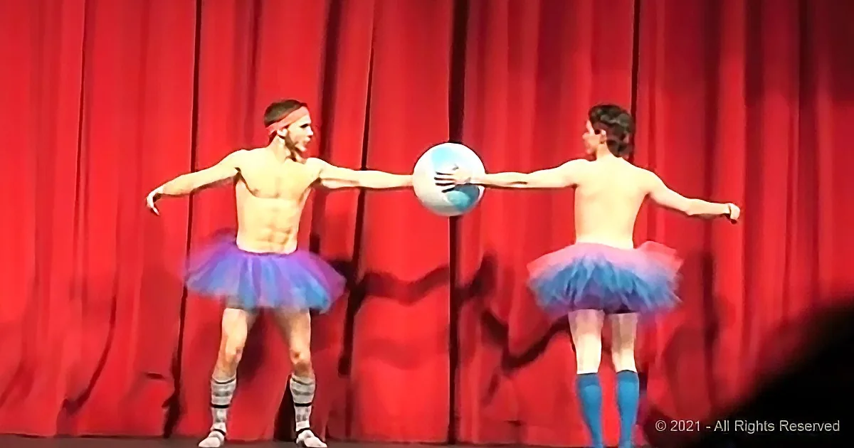 Two shirtless men in tutus perform a synchronized dance with a ball against a red curtain backdrop.