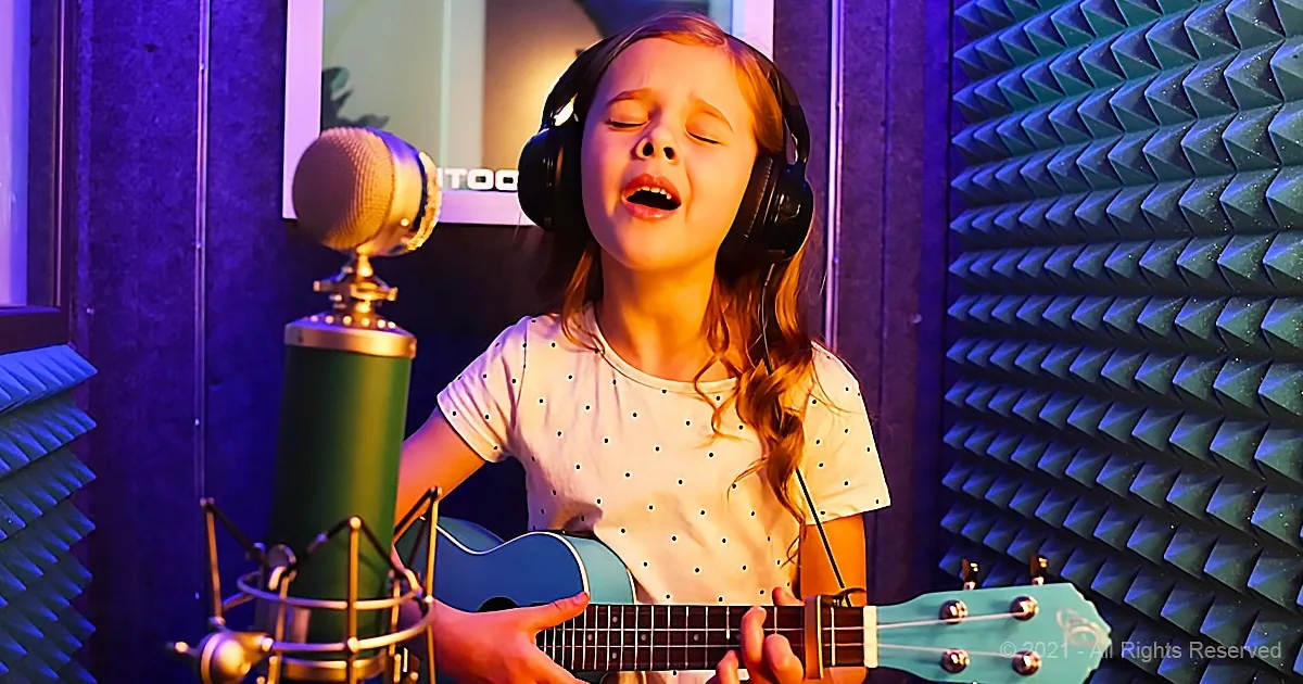 A young girl sings into a microphone while playing a ukulele in a recording studio.