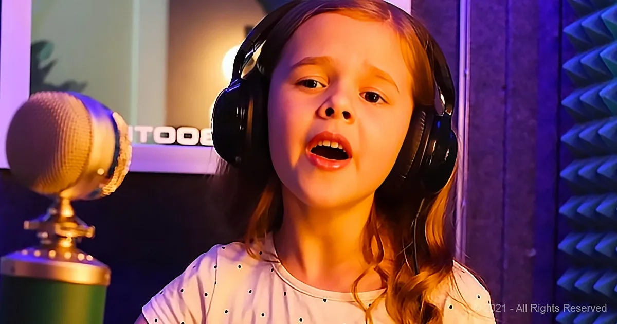 A young girl wearing headphones sings into a microphone in a recording studio with purple lighting.