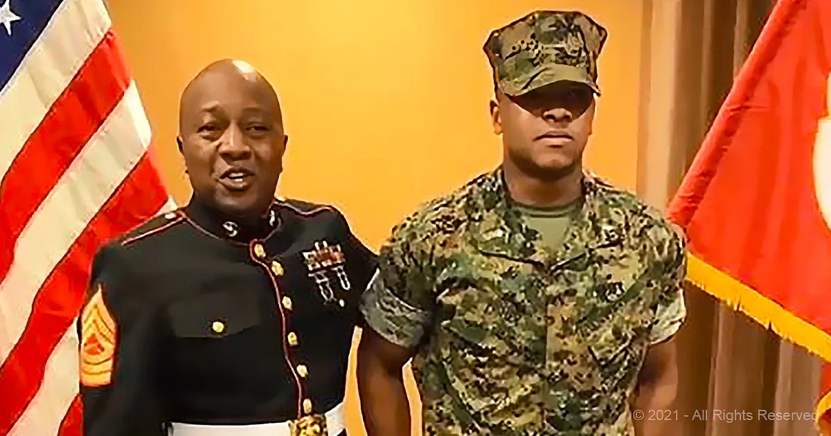 A man in a military uniform stands next to another in camouflage, both in front of American flags.