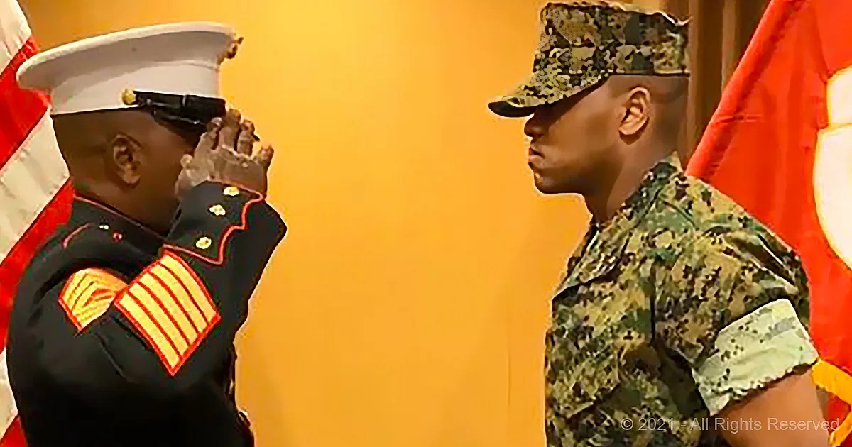 A Marine in uniform salutes another in camouflage, both in front of flags, under warm lighting.