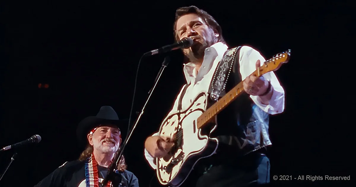 A man in a white shirt and black vest plays guitar on stage, singing into a microphone.