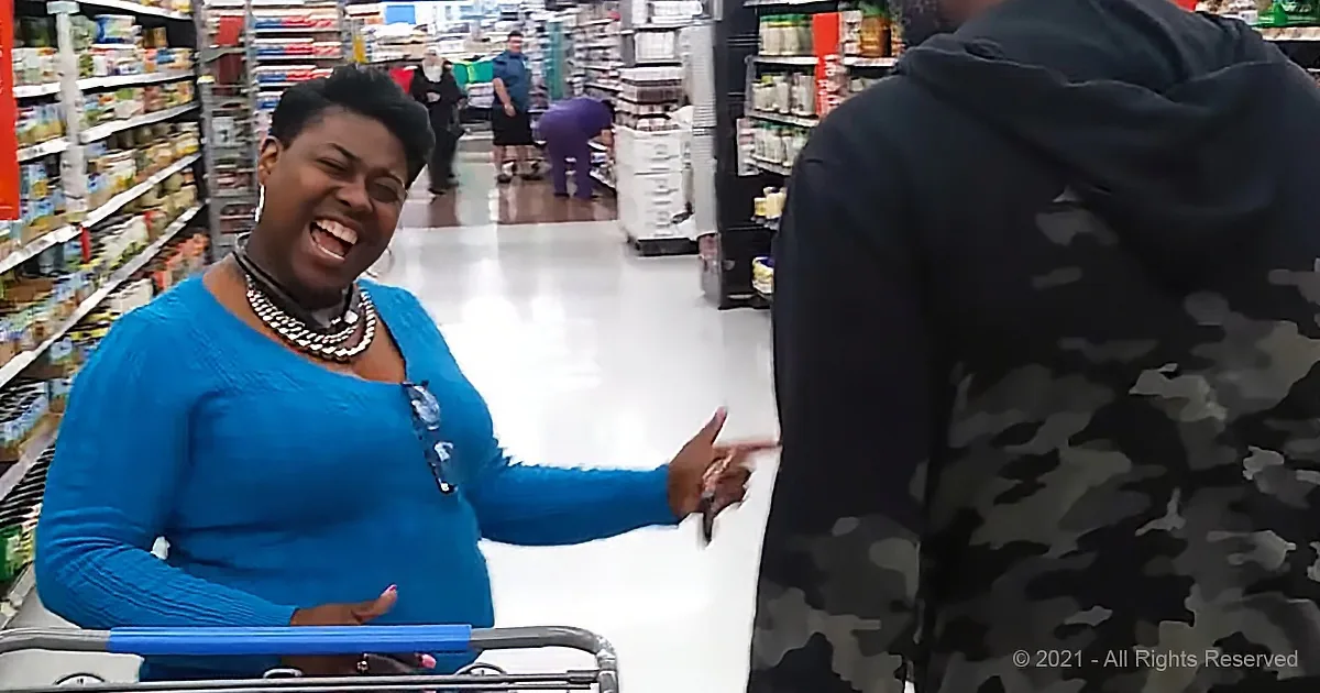 A woman in a blue sweater laughs while holding a shopping cart in a grocery store aisle.
