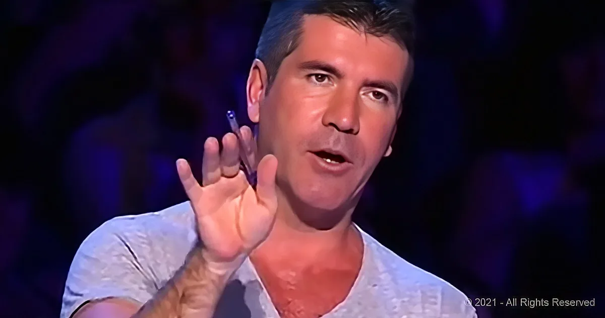 Simon Cowell gestures with his right hand, wearing a white shirt, in a dimly lit setting.