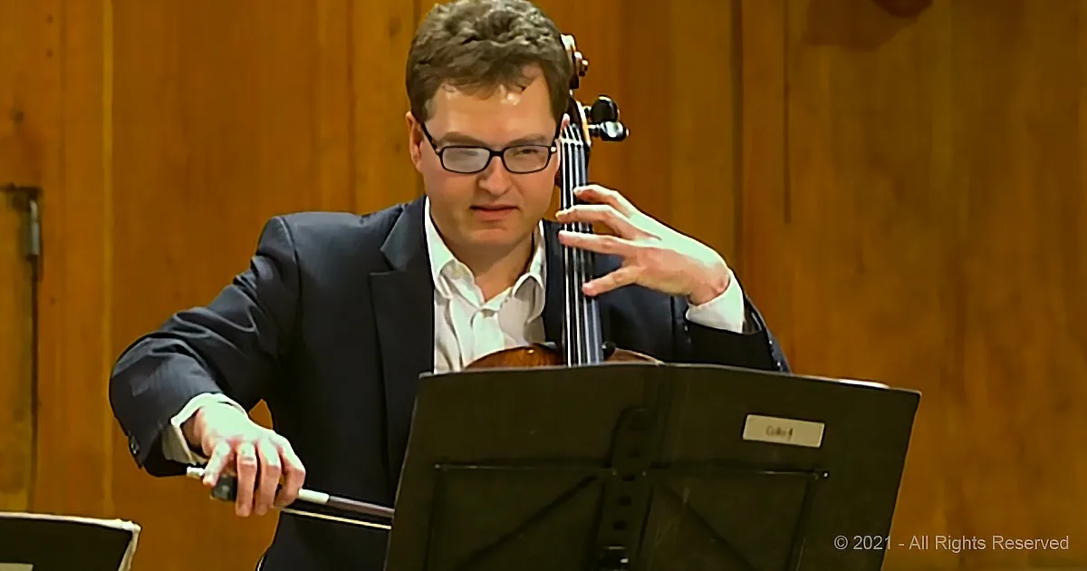 A man in a suit plays a cello, holding the bow and looking at sheet music.