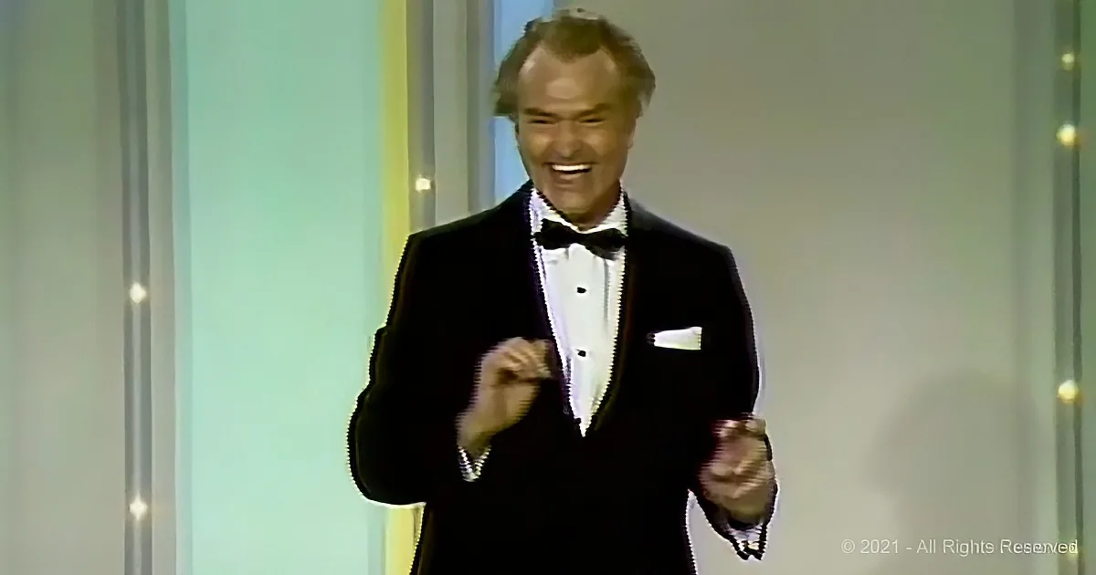 A man in a tuxedo smiles while holding a microphone on a stage.