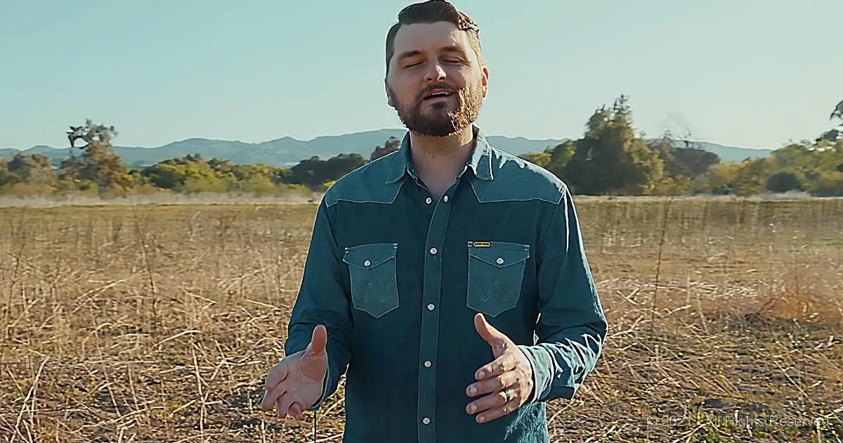 A man in a teal shirt stands in a field with mountains and trees in the background.