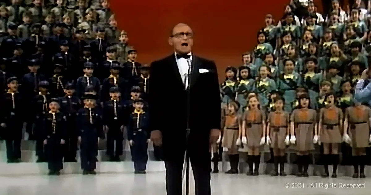 A man in a tuxedo stands in front of a choir, singing, with a large audience in the background.