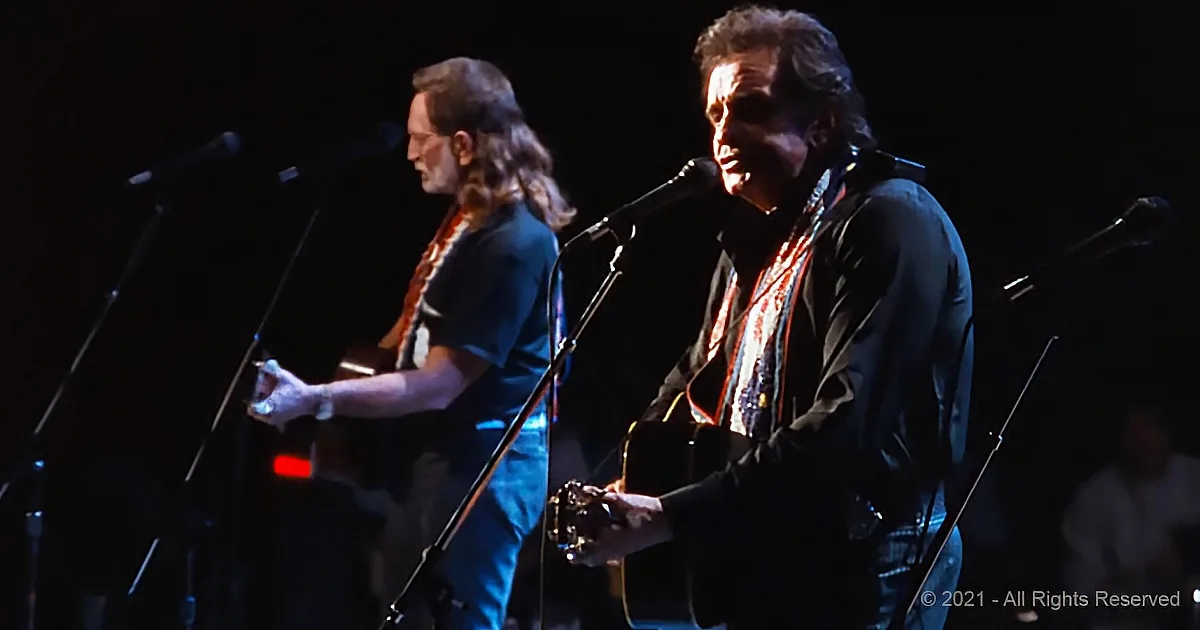 A man in a black shirt and jeans plays guitar on stage with a microphone, another musician in the background.