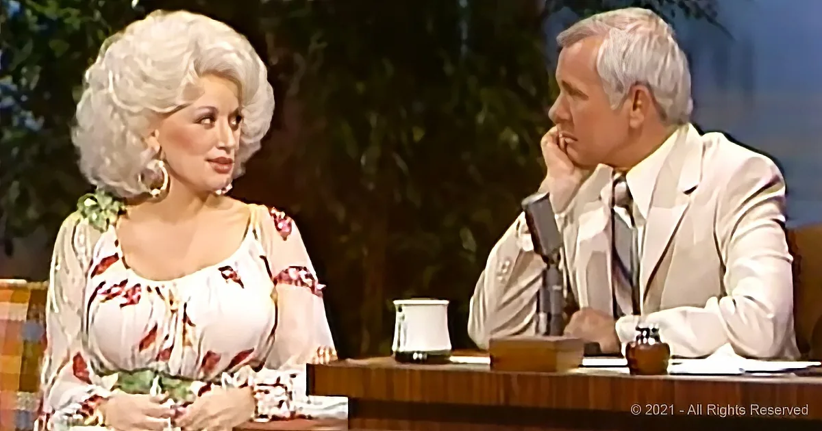 Dolly Parton in floral dress talks to man in white suit on talk show set.