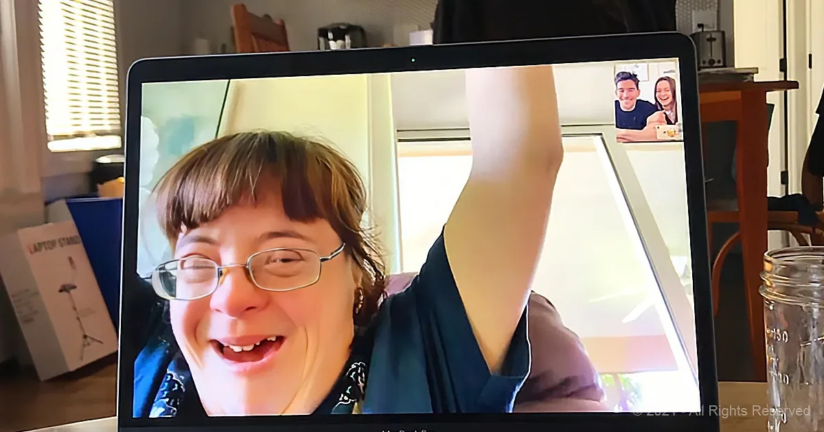 A woman with glasses raises her arm, smiling, in a video call on a laptop screen.