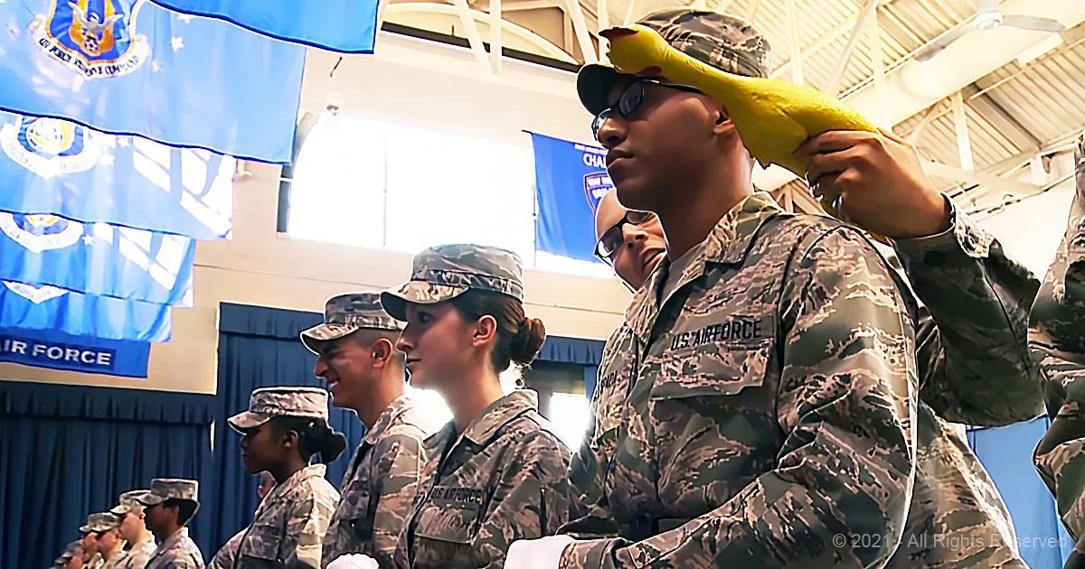 A soldier in camouflage holds a yellow rubber duck, surrounded by other uniformed personnel in a formal setting.