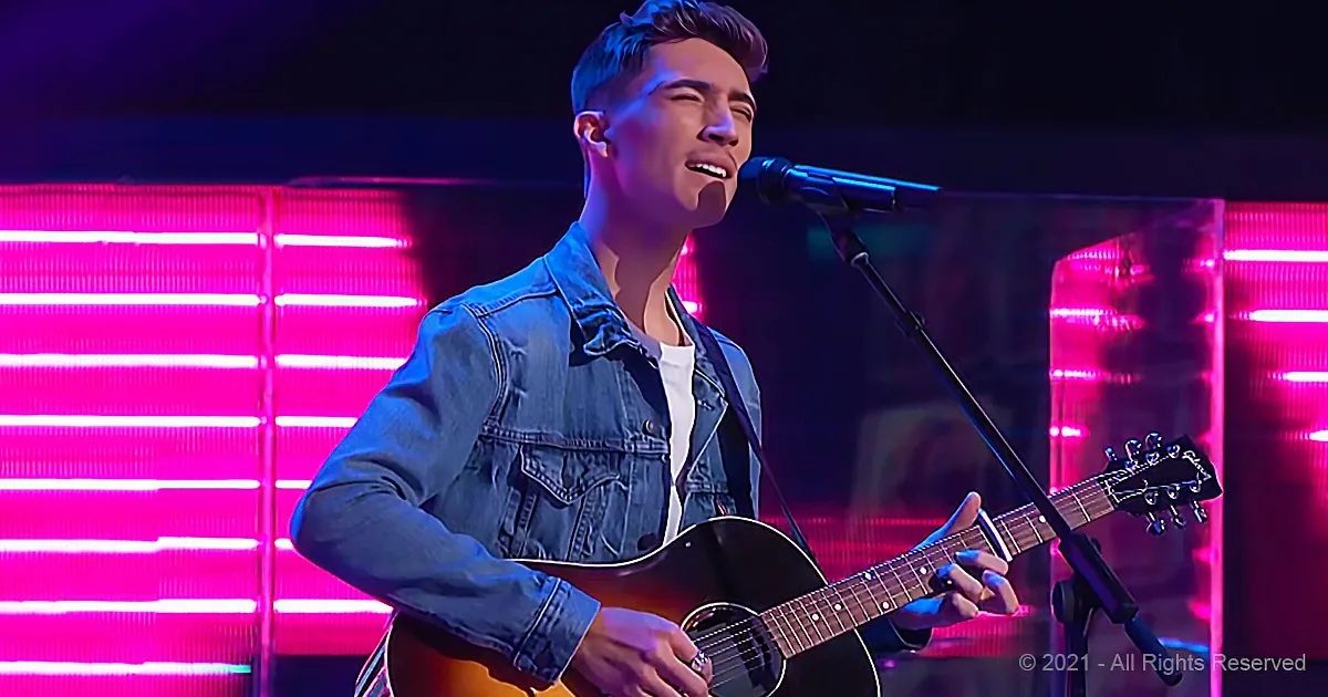A man in a denim jacket plays an acoustic guitar on stage with pink lights behind him.