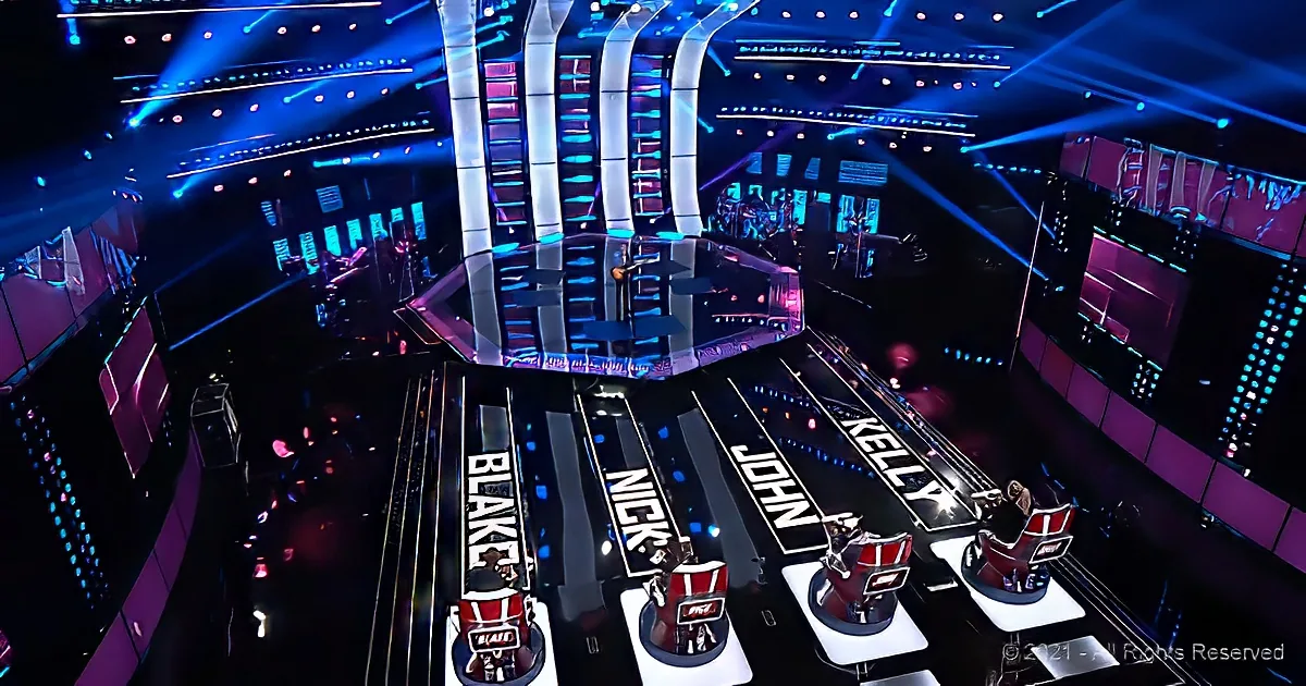 A stage with judges' chairs labeled Blake, Nick, John, and Kelly, illuminated by vibrant blue and purple lights.