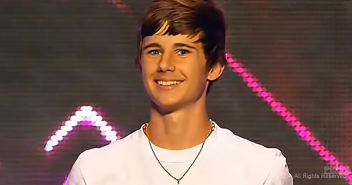 A smiling young man in a white shirt and gold necklace stands in front of a pink and white light display.