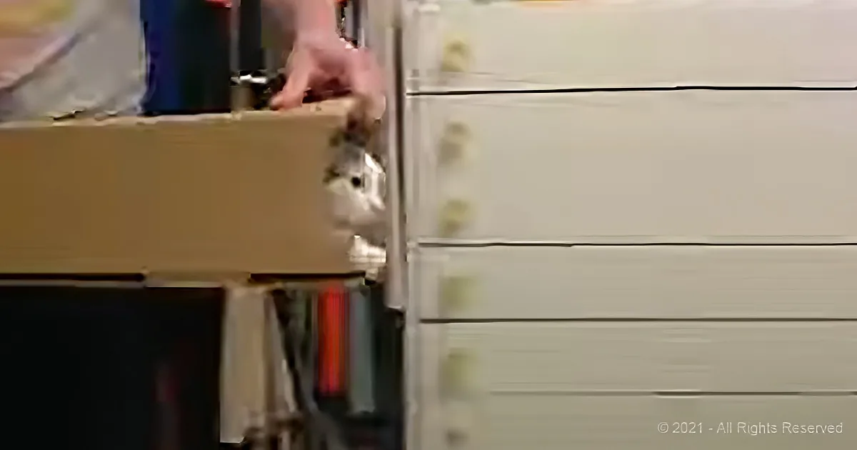 A hand reaches into a cardboard box on a shelf, with a white dresser and books in the background.