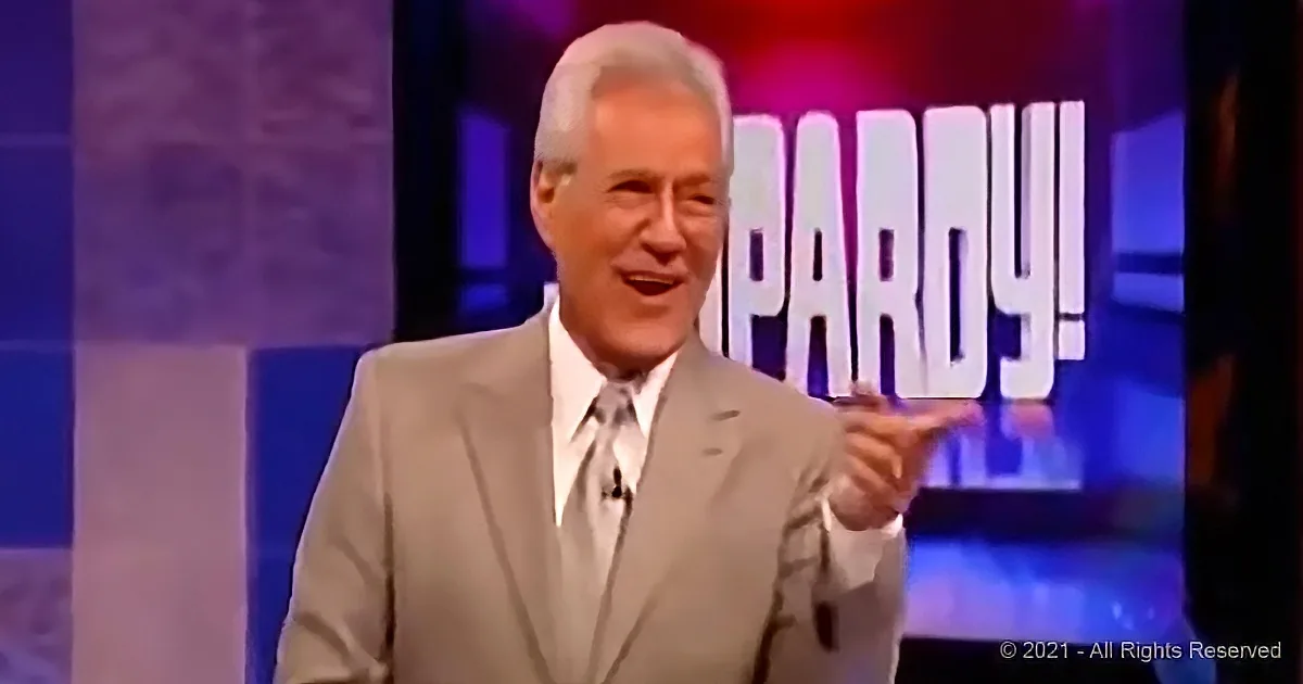 A man in a suit points to the right on a game show set with "Jeopardy!" in the background.
