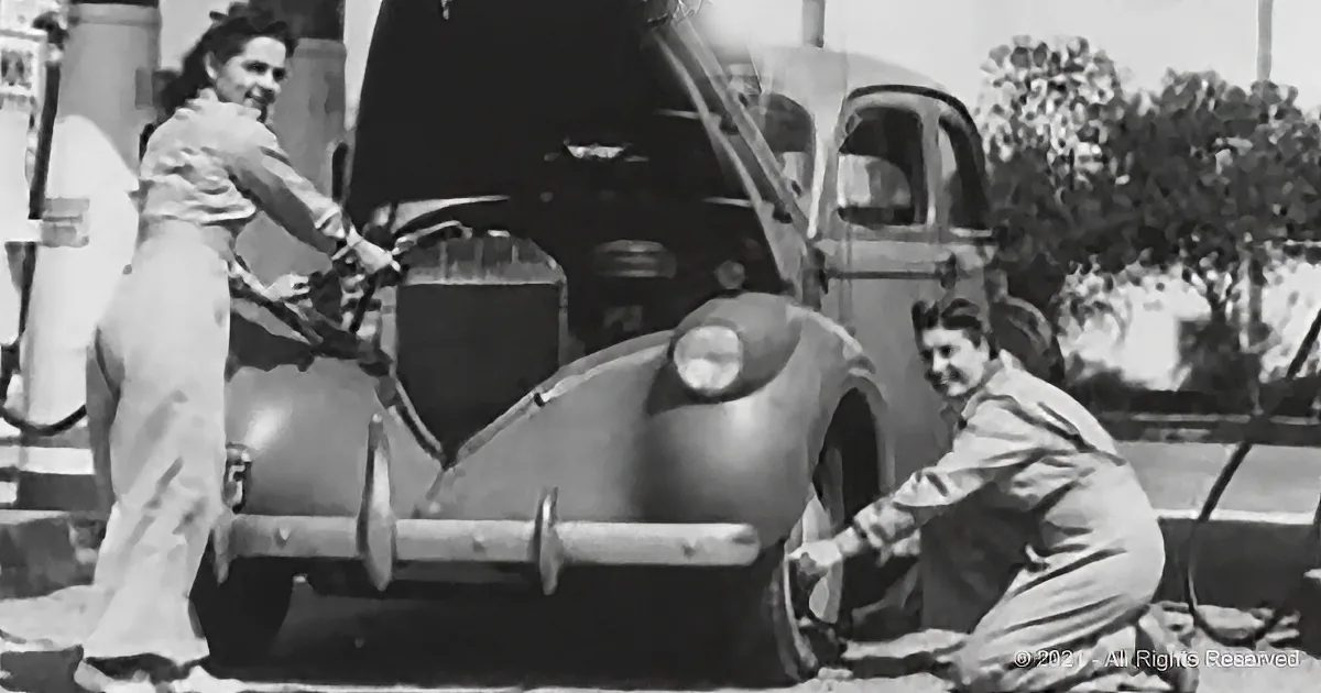 A woman in coveralls pumps gas into a vintage car while another person works on the tire.