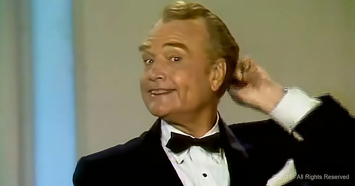 A man in a tuxedo adjusts his hair while smiling, possibly on a TV show.
