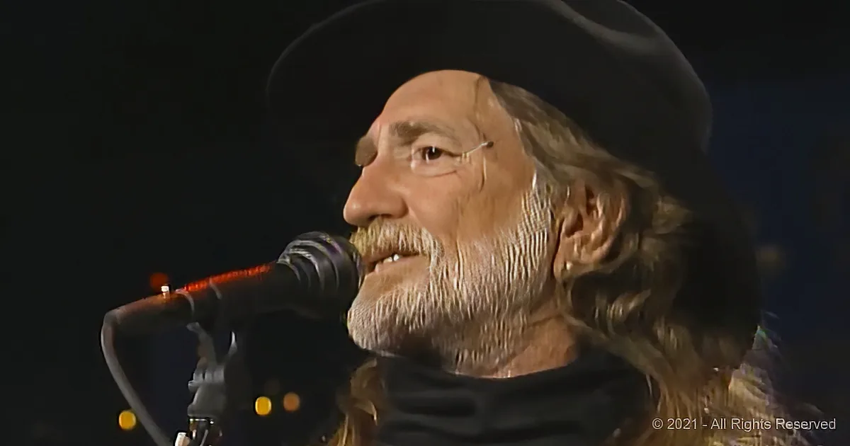 A man with long hair and a beard sings into a microphone, wearing a dark hat.