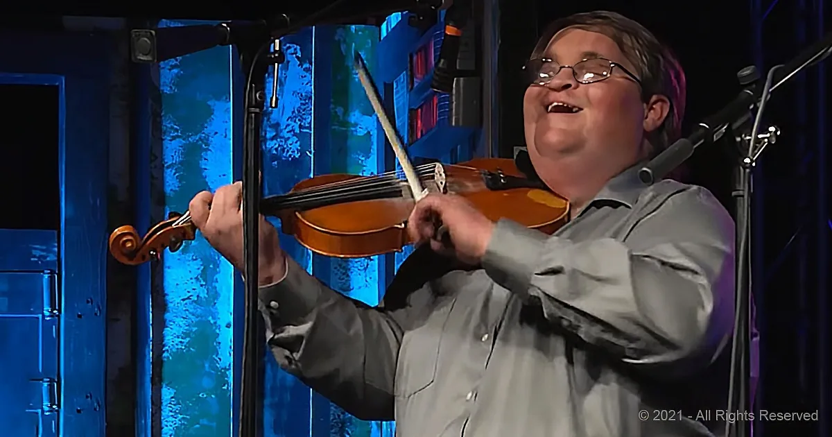 A man in a gray shirt plays a violin on stage with a microphone and blue lighting.