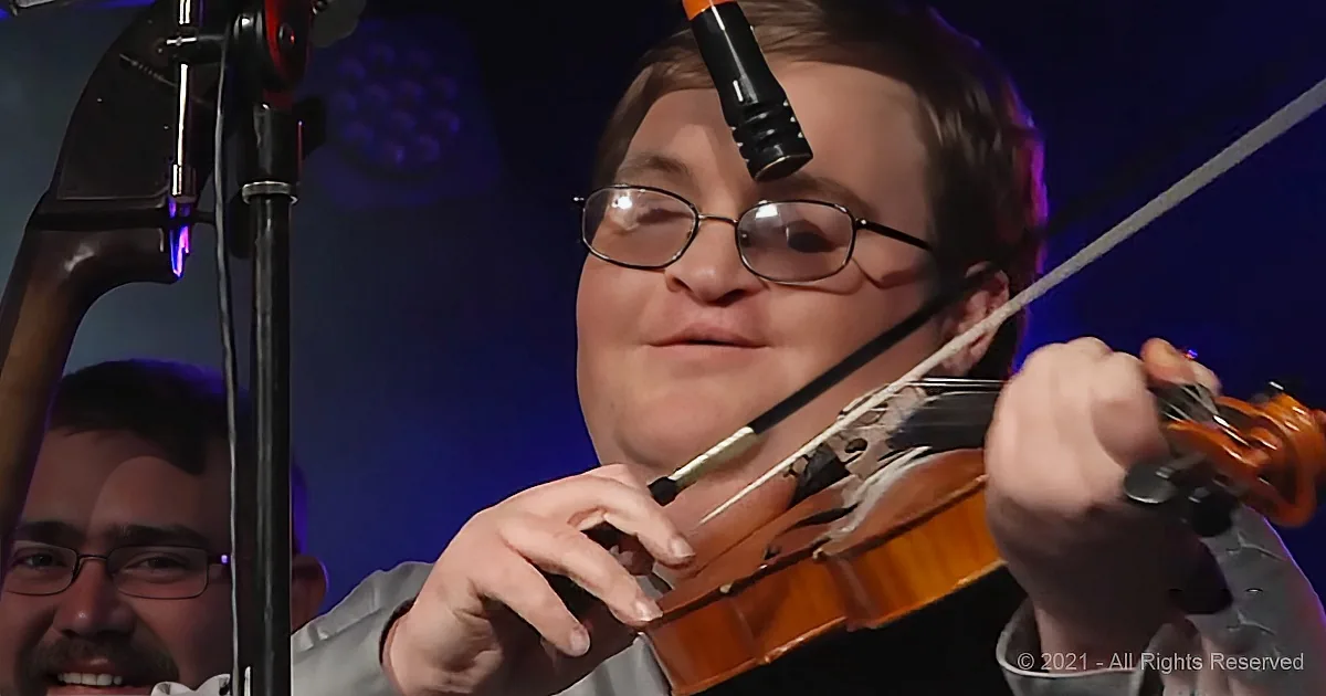 A person wearing glasses plays a violin with a microphone above, while another person smiles in the background.