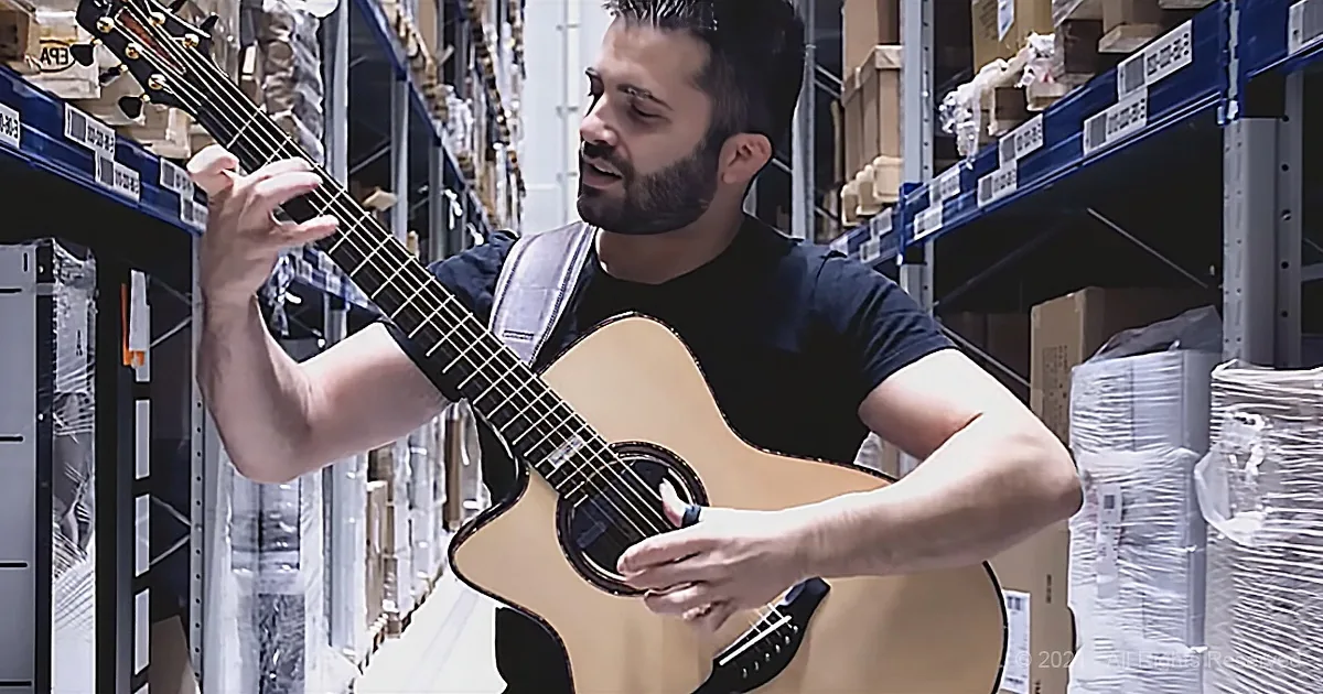 A man with a beard plays an acoustic guitar in a warehouse aisle.