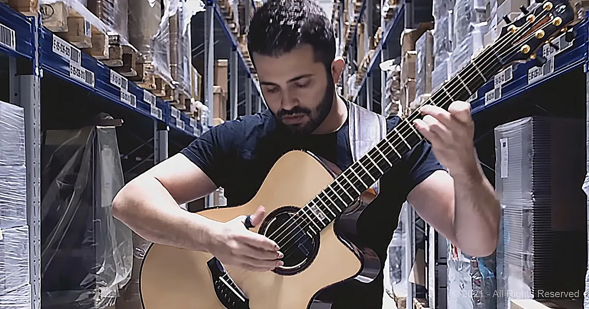 A man plays an acoustic guitar in a warehouse aisle, surrounded by shelves and boxes.