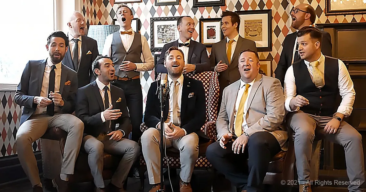 A group of men in formal attire sing together, some seated and some standing, in a room with patterned wallpaper.