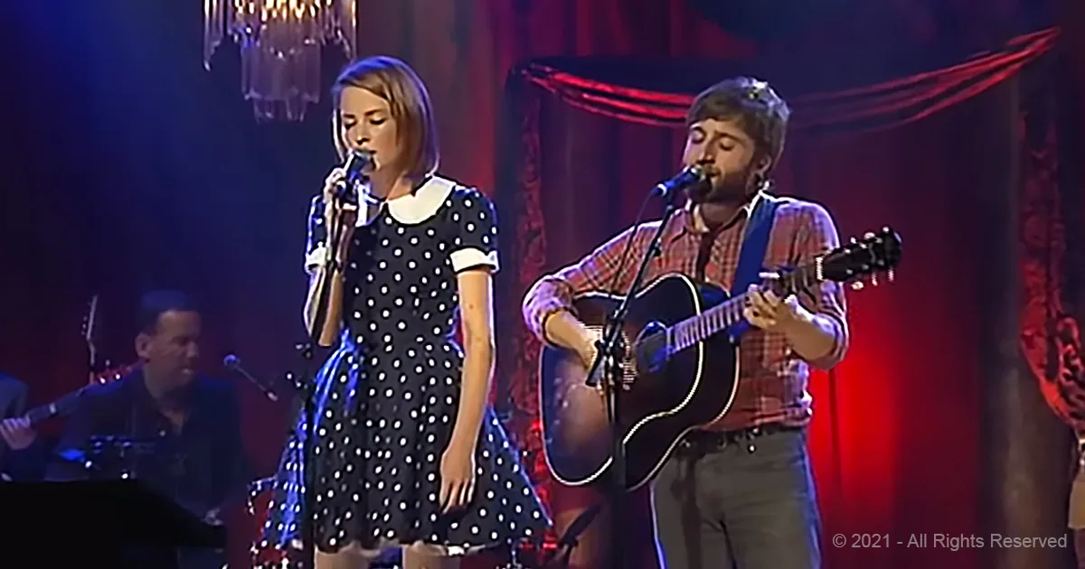 A woman in a polka-dot dress sings into a microphone, accompanied by a man playing guitar on stage.