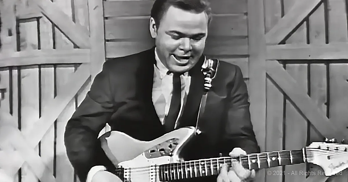 A man in a suit plays a white electric guitar, smiling, against a wooden backdrop.