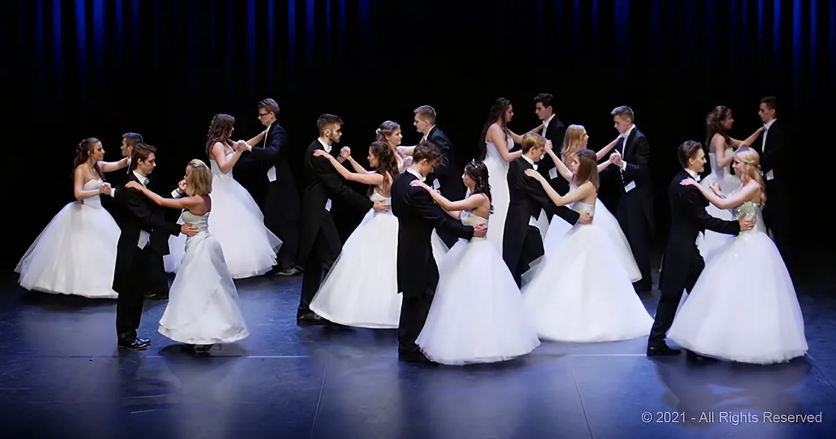 A group of elegantly dressed dancers in formal attire perform a synchronized dance on a stage.
