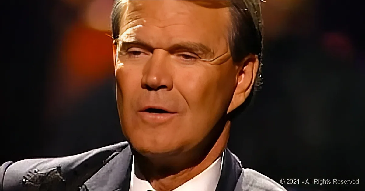 A man in a suit and tie is speaking, with a blurred background and some text in the corner.