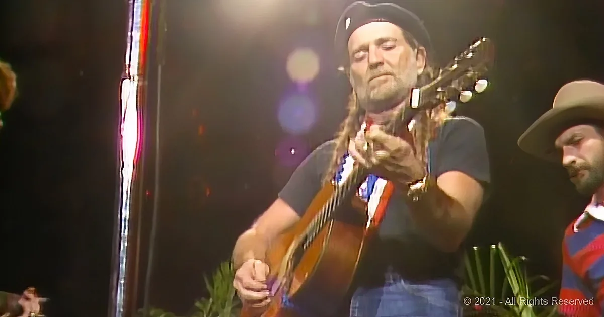 A man with long hair and a hat plays a guitar on stage, surrounded by other musicians.