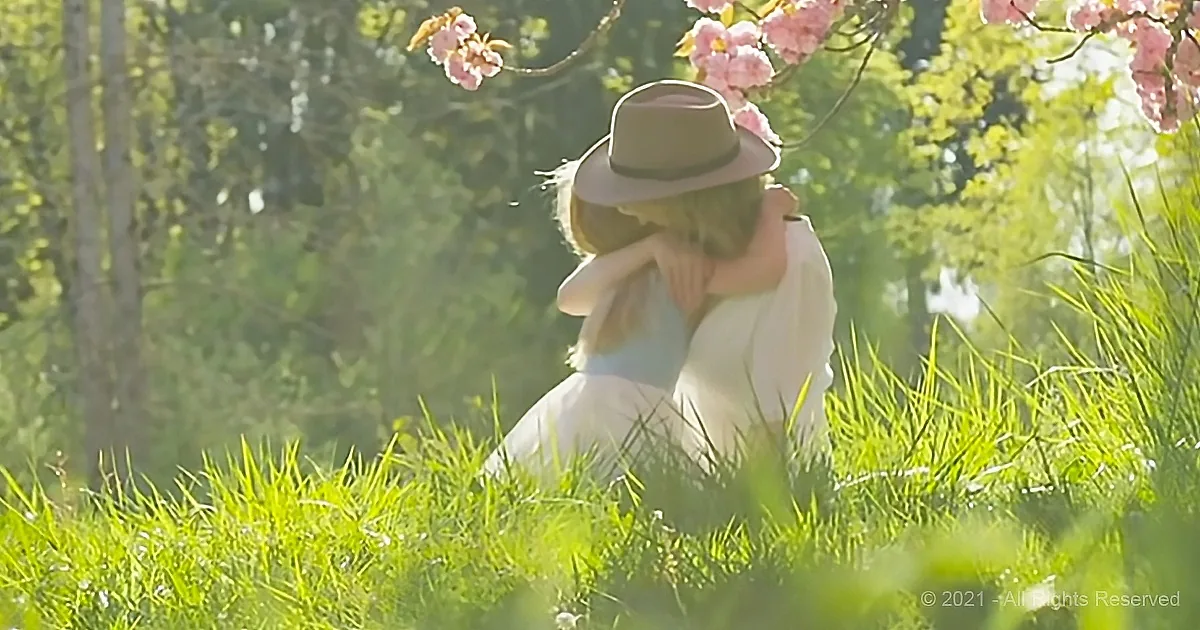 A person in a white dress and hat sits in a grassy field, surrounded by blooming pink flowers and greenery.