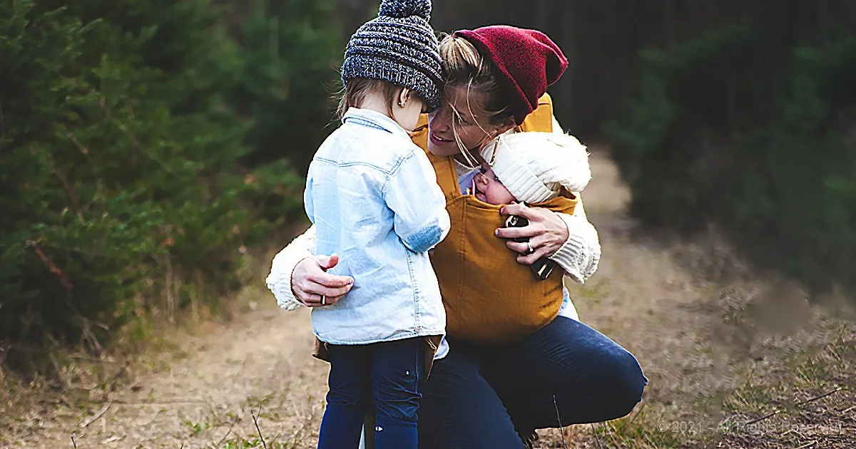 A woman hugs a child and carries another in a sling, both wearing winter hats, in a forest setting.